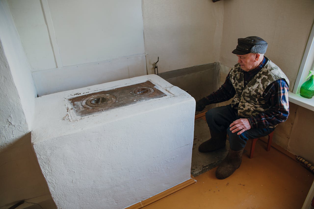 Elderly man wearing warm clothes, sitting by a vintage stove inside a cozy room.